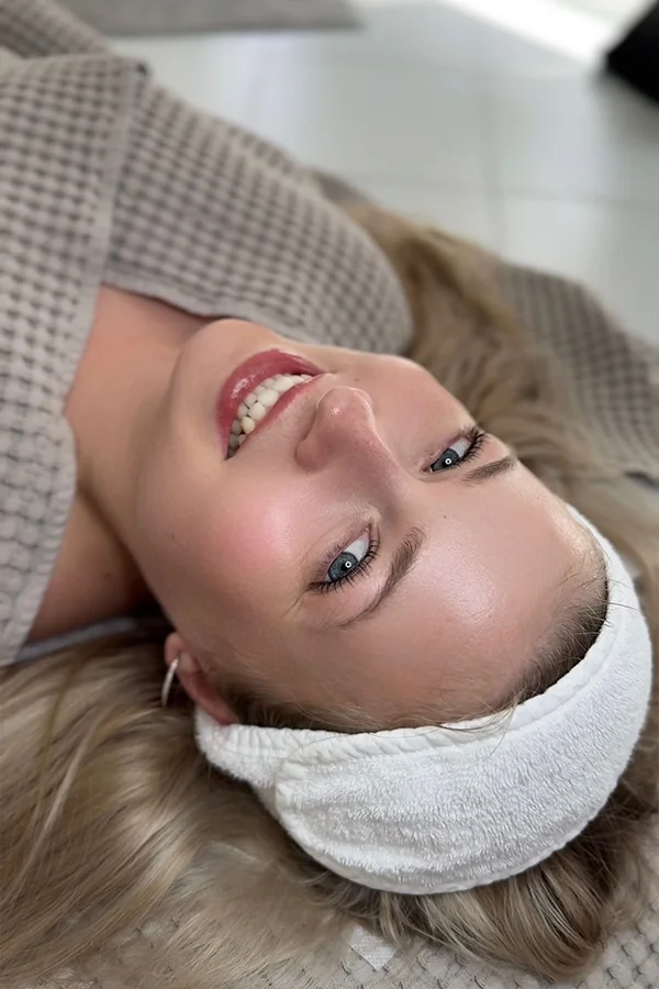 Eine Frau mit langen blonden Haaren liegt auf einer strukturierten Oberfläche und lächelt in die Kamera. Sie trägt ein weißes Stirnband und einen beigen Bademantel mit Waffelmuster, was eine entspannte und gemütliche Atmosphäre schafft.
