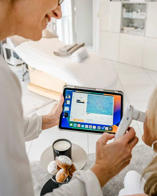 Eine Person hält ein Tablet mit einer Hautanalyse-Software in der Hand, während sie das Gesicht einer anderen Person mit einem Handgerät untersucht. In der Nähe stehen auf einem Tisch eine Tasse und ein Teller mit Gebäck. Die Umgebung scheint eine moderne Klinik oder ein Spa zu sein.