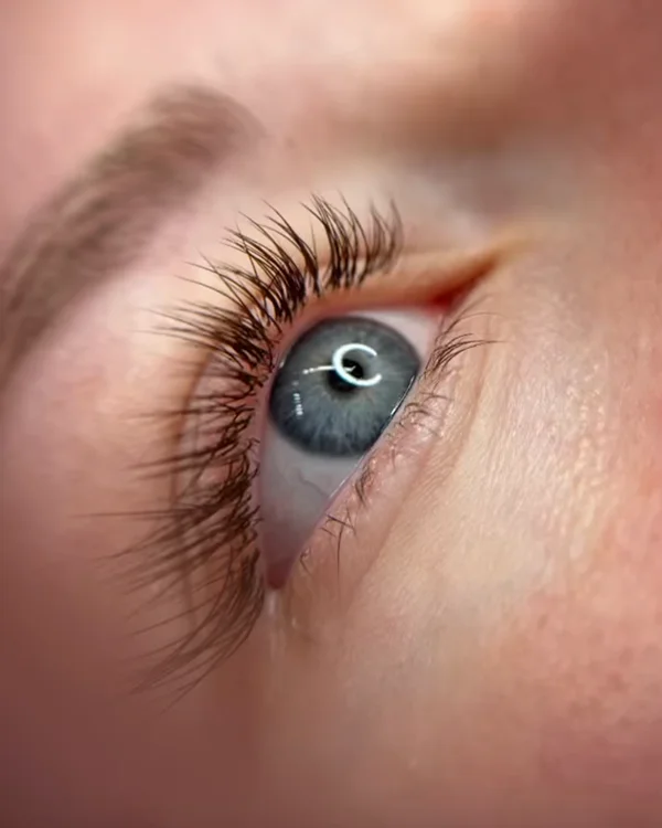 Eine Nahaufnahme eines blauen Auges, das nach oben blickt und detaillierte Wimpern, Augenbrauen und die Struktur der Haut um das Auge herum zeigt. In der Pupille ist eine Ringlichtreflexion sichtbar.
